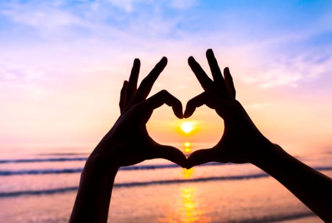 Two hands are meeting to form a heart symbol with their fingers against a rainbow sunset